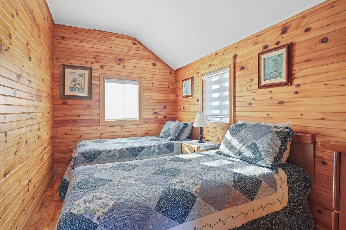 Ein gemütliches Blockhauskabinenzimmer mit Holzwänden, zwei Betten mit Patchworkdecken, einem Fenster mit Jalousien und gerahmten Bildern an den Wänden.