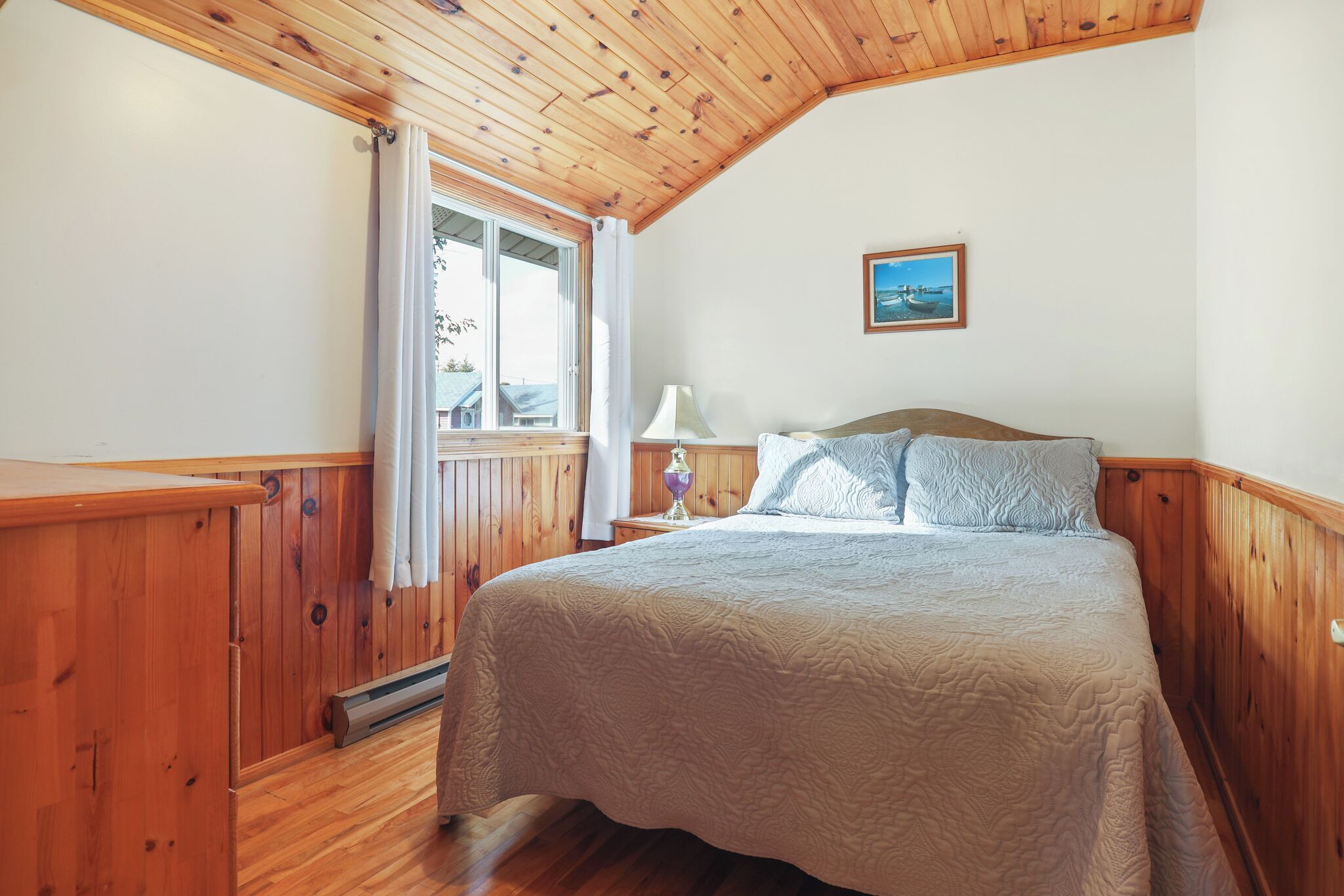 Une chambre avec un plafond en bois, un lit recouvert d’une courtepointe, une table de chevet avec une lampe et une fenêtre avec des rideaux.