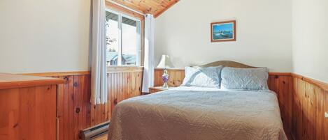 Une chambre avec un plafond en bois, un lit recouvert d’une courtepointe, une table de chevet avec une lampe et une fenêtre avec des rideaux.