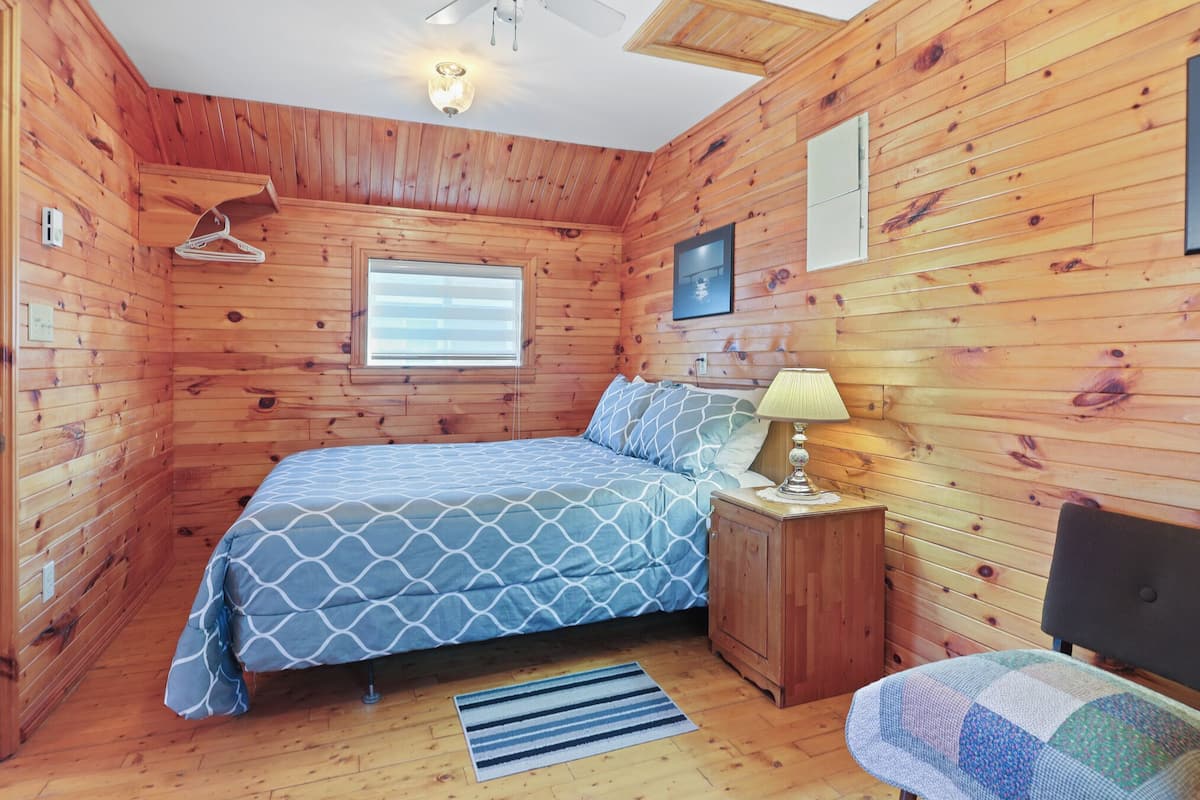 Ein Zimmer mit Holzwänden, einem Bett, einem Nachttisch, einem Kleiderständer, einem Fenster und einem an der Wand hängenden Bild.