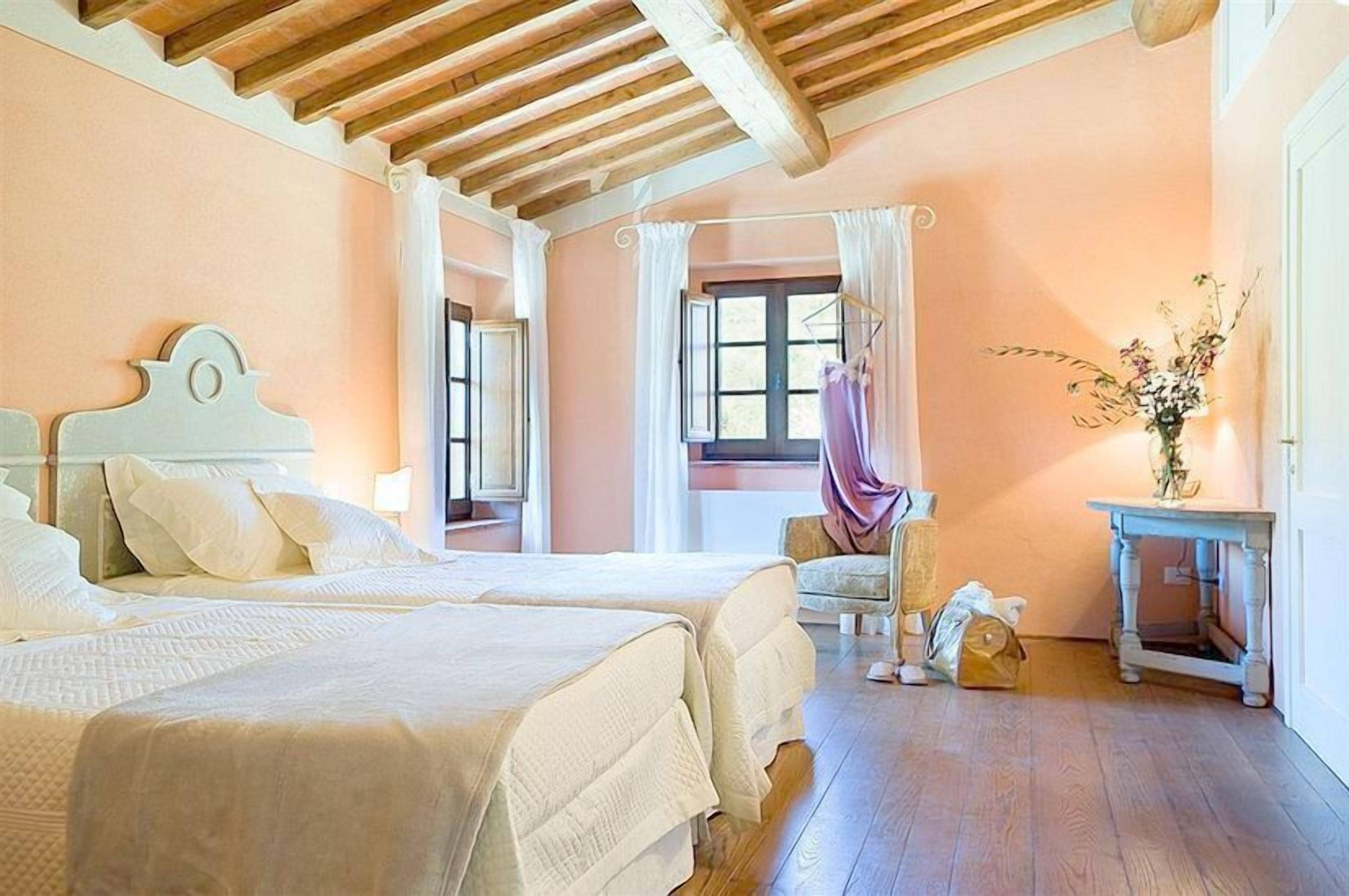 A bedroom with two beds, wooden ceiling beams, a window with curtains, a chair, and a small table with a vase of flowers.