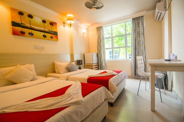 A hotel room with two beds, a desk, a chair, a window with curtains, an air conditioner, and a painting on the wall.