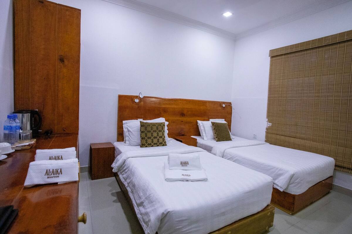 Habitación de hotel con dos camas individuales, un cabecero de madera, un escritorio de madera con una tetera y una ventana con persiana.