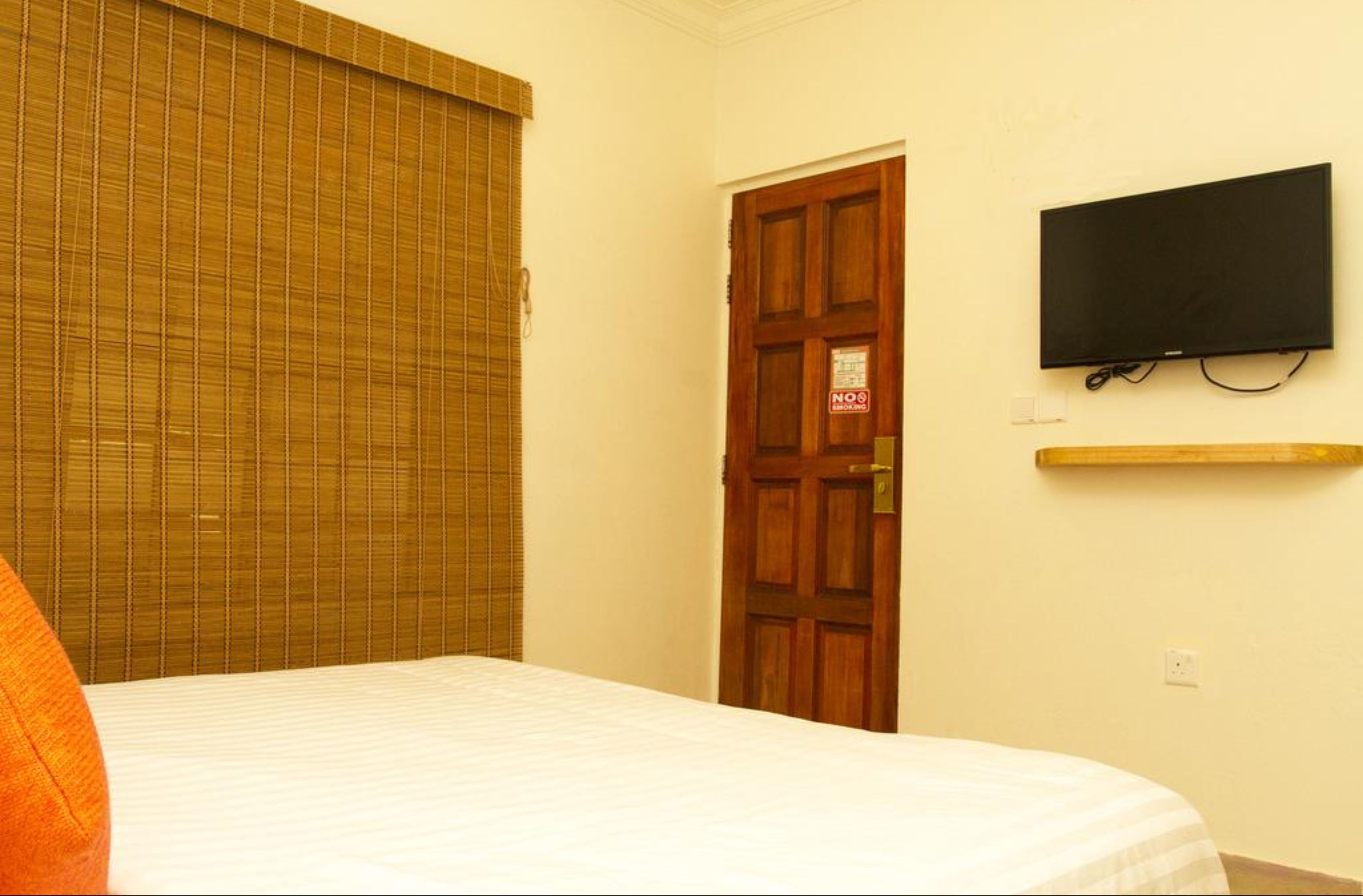 A hotel room with a bed, a closed door, a television mounted on the wall, and a window with blinds.