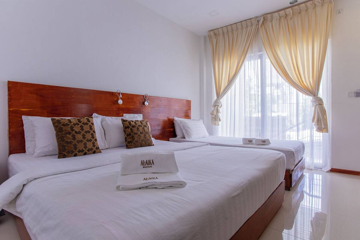 Habitación de hotel con cabecera de madera, dos camas, ropa de cama blanca, almohadas estampadas y una ventana con cortinas translúcidas.