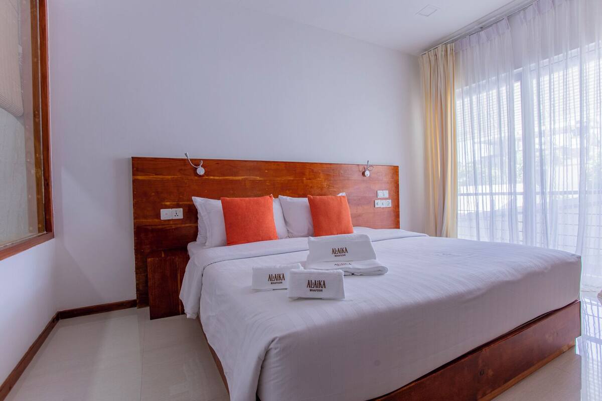 Una cama bien tendida con sábanas blancas y almohadas naranjas, un cabecero de madera y una ventana con cortinas de encaje.