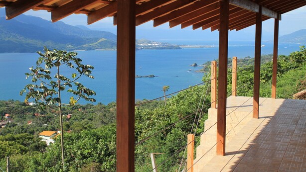 Une pergola en bois, dotée d’un sol carrelé, offre une vue sur un paysage côtier avec des montagnes et une étendue d’eau.