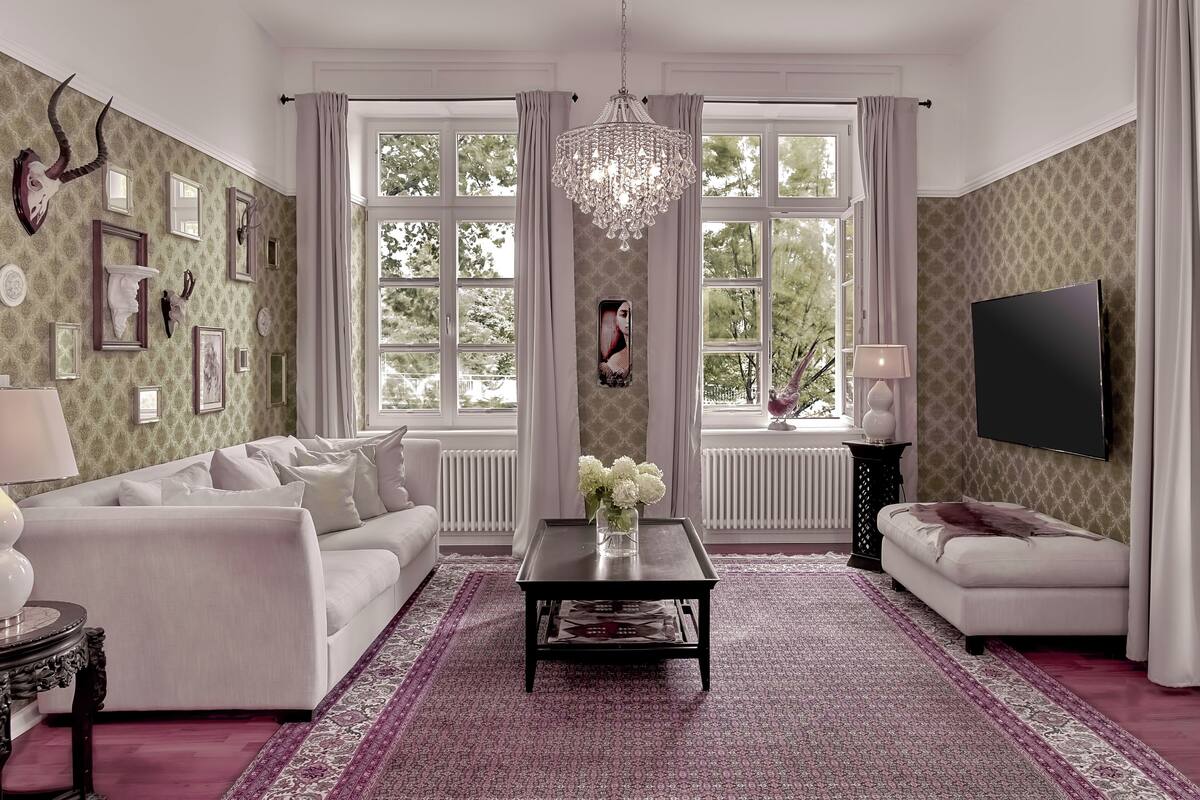 Ein geräumiges Wohnzimmer mit einem weißen Sofa, einem Couchtisch mit einer Vase, einem großen Fenster mit Vorhängen und einer Chandelier.