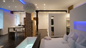 A modern bedroom with a large bed, a glass-enclosed bathtub, a brick accent wall, and a desk with chairs. - Boutique Suites and Residences Heidelberg – Alte Zigarrenmanufaktur (Heidelberg)