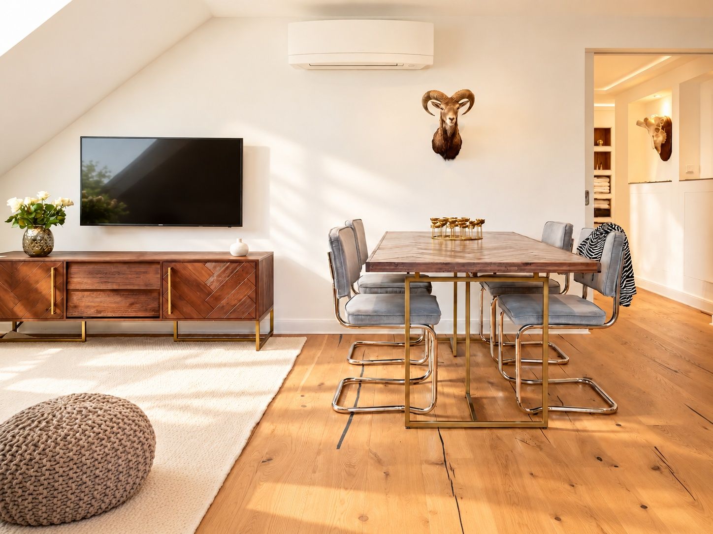 A modern dining area with a wooden table and metal chairs, a mounted television, and a decorative ram skull on the wall.