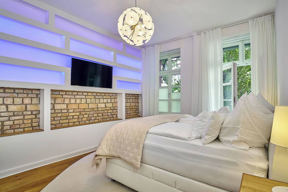 Ein Schlafzimmer mit an einer Ziegelwand montiertem Fernseher, einem Bett mit weißer Bettwäsche und einem Fenster mit Vorhängen.