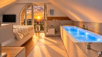 A modern bedroom with a large bed, a bathtub, and a balcony with a sunset view.