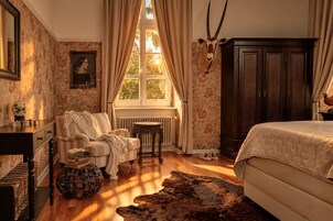Una habitación con cama, una silla, un radiador, una ventana con cortinas y una cabeza de animal colgada en la pared.