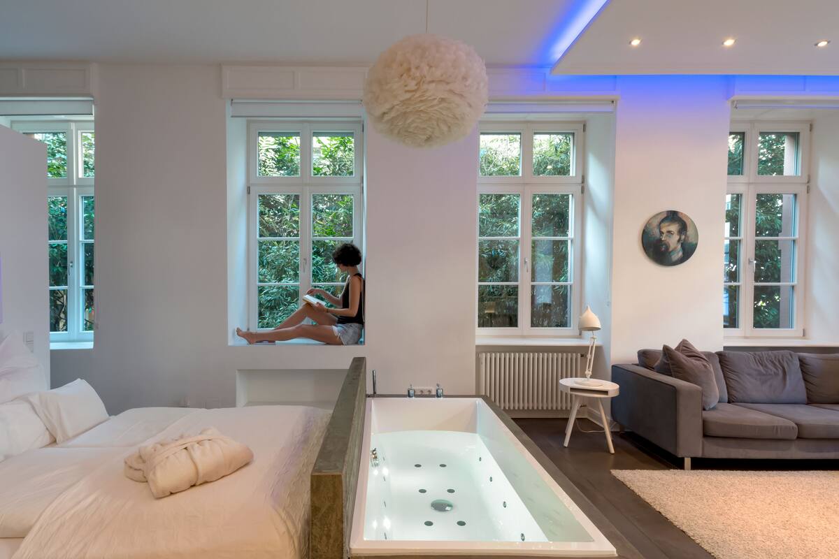 Ein modernes Schlafzimmer mit einem großen Bett, einer Badewanne, einer Sitzbank an einem Fenster und einem grauen Sofa.