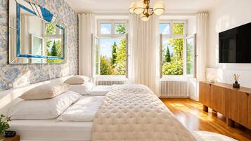 Quarto com cama grande, televisão, radiador e vista para árvores pelas janelas.