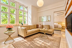 Een moderne woonkamer met een beige bank, een bijzettafeltje, een groot raam met uitzicht op groen en een flatscreen televisie.