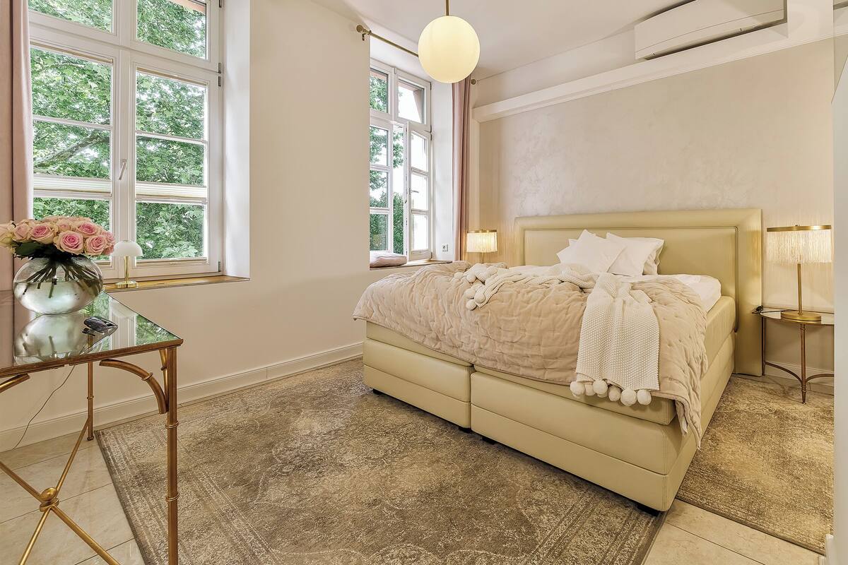 Ein Schlafzimmer mit großem Fenster, einem Bett mit weißem Kopfteil und einem Nachttisch mit Lampe.