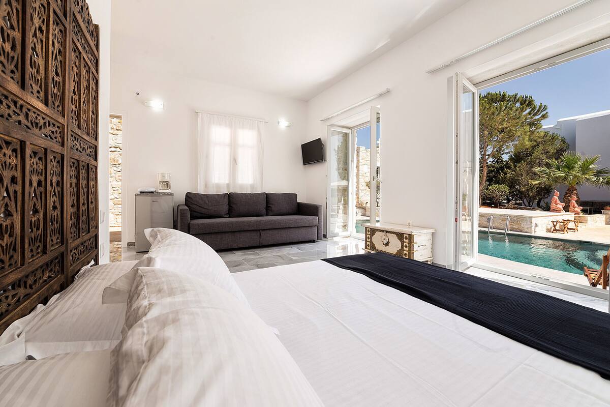 A bedroom with a large bed, a grey sofa, a wooden decorative panel, a TV, and a view of an outdoor swimming pool.