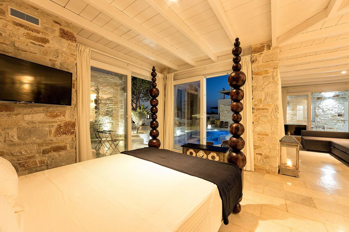 A bedroom with a large bed, stone walls, a TV, and a view of a pool.