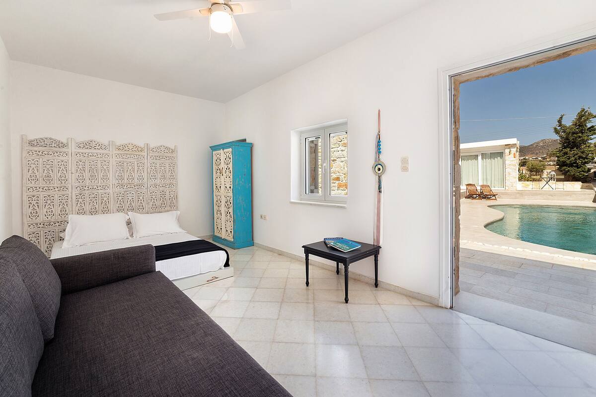 A modern bedroom with a bed, a grey sofa, a small table, and a view of a swimming pool.