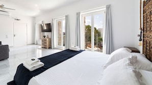 A modern hotel room with a large bed, a flat-screen TV, a wooden headboard, and a balcony with a view. - Villa Pela (Paros)