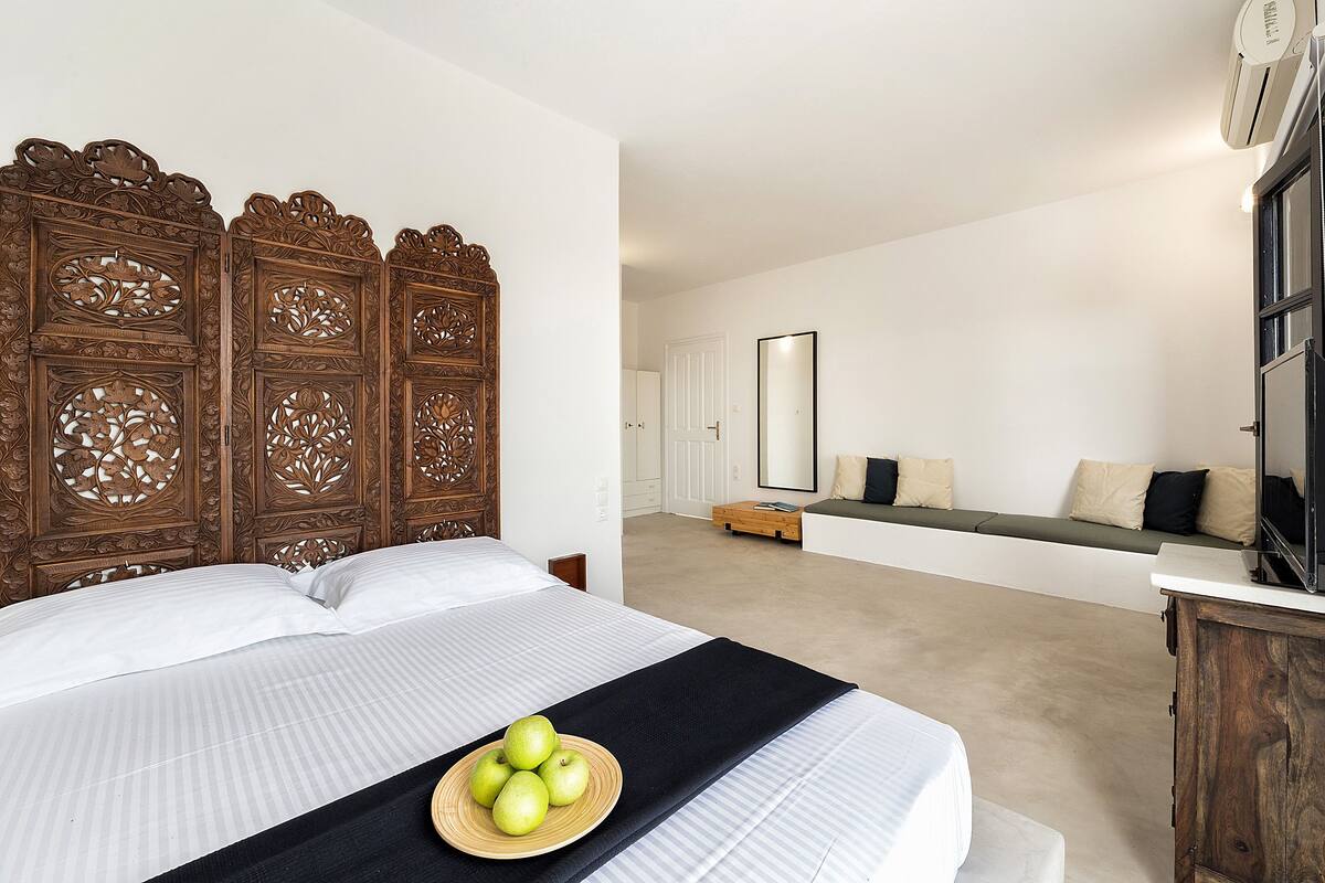 A bedroom with a large bed, a wooden headboard, a bedside table, and a seating area with cushions.