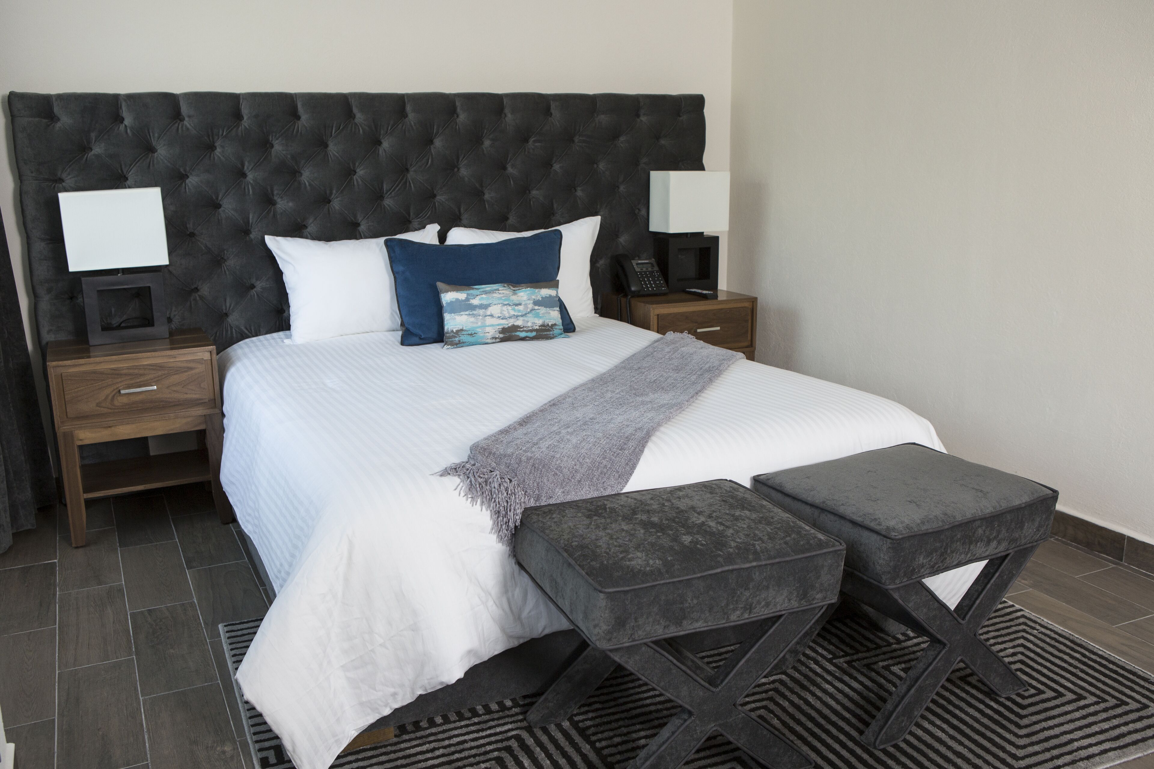 A neatly arranged bedroom with a tufted headboard, a bed with white bedding and blue pillows, a bedside table with a lamp, and a small stool.