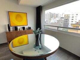 Un salon moderne avec une table ronde en verre, un meuble en bois, une grande œuvre d’art jaune et une vue sur les bâtiments par la fenêtre.