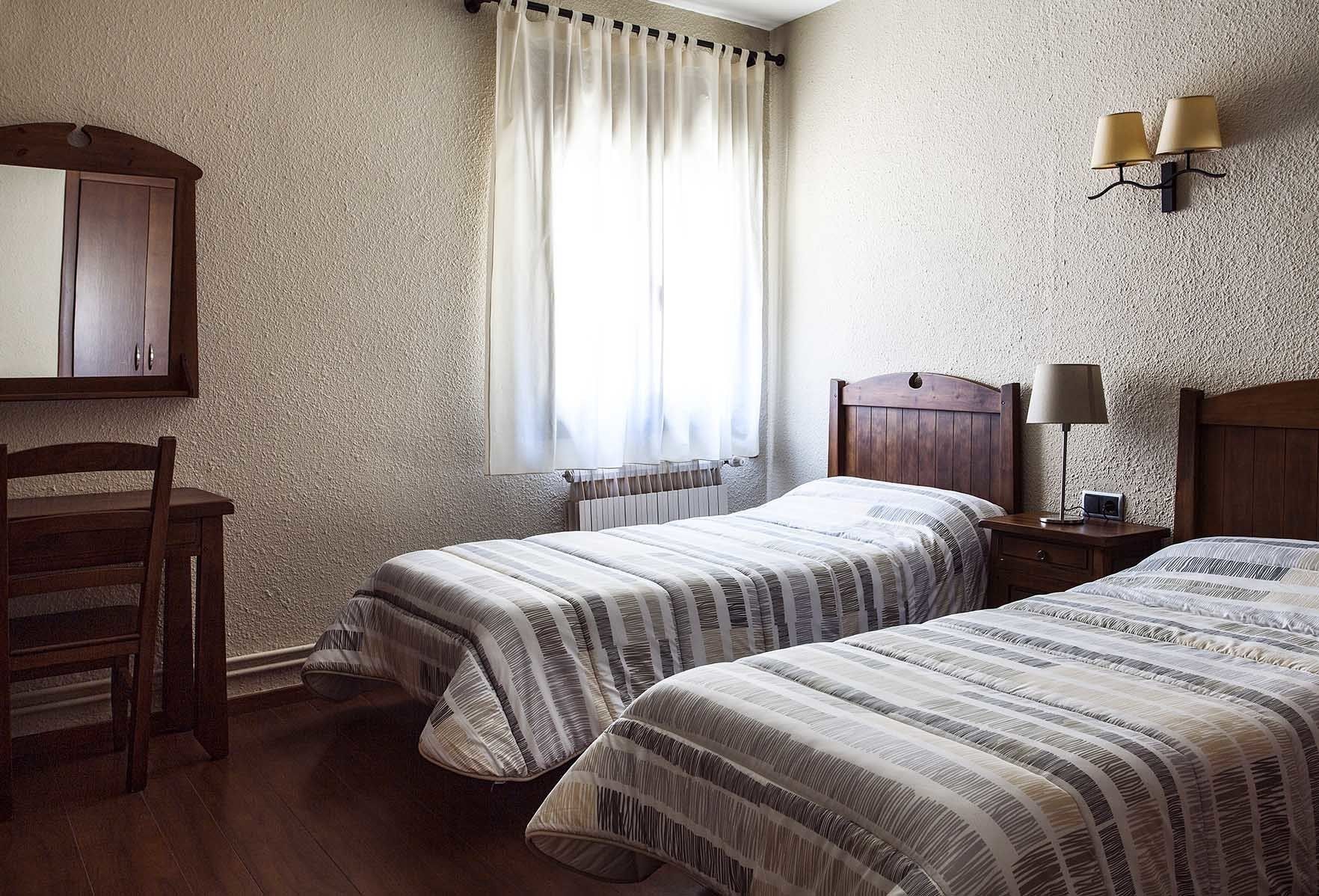 Un dormitorio con dos camas, una silla de madera, una mesita de noche con lámpara, un estante en la pared y una ventana con cortinas.
