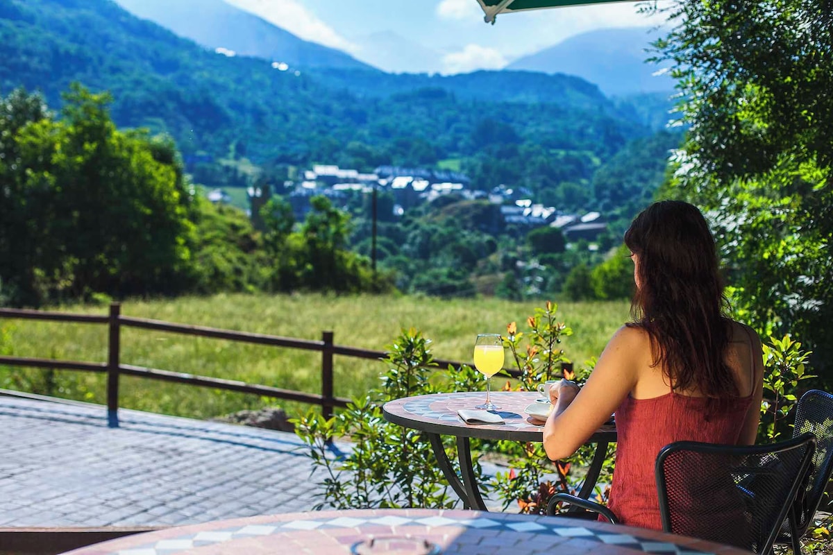 A person seated at an outdoor table with a glass of drink, overlooking a scenic view of a valley and mountains.