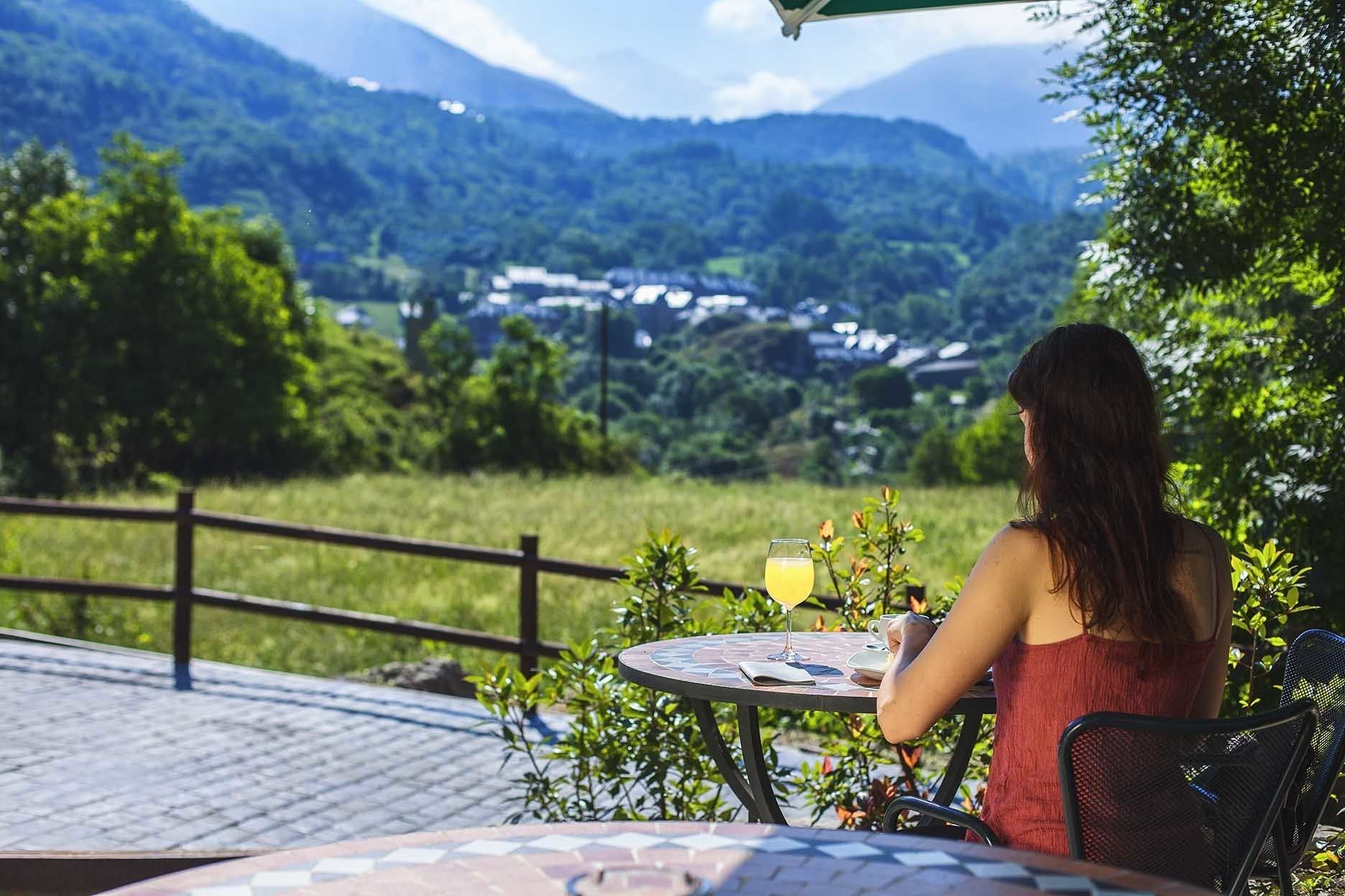 A person seated at an outdoor table with a glass of drink, overlooking a scenic view of a valley and mountains.