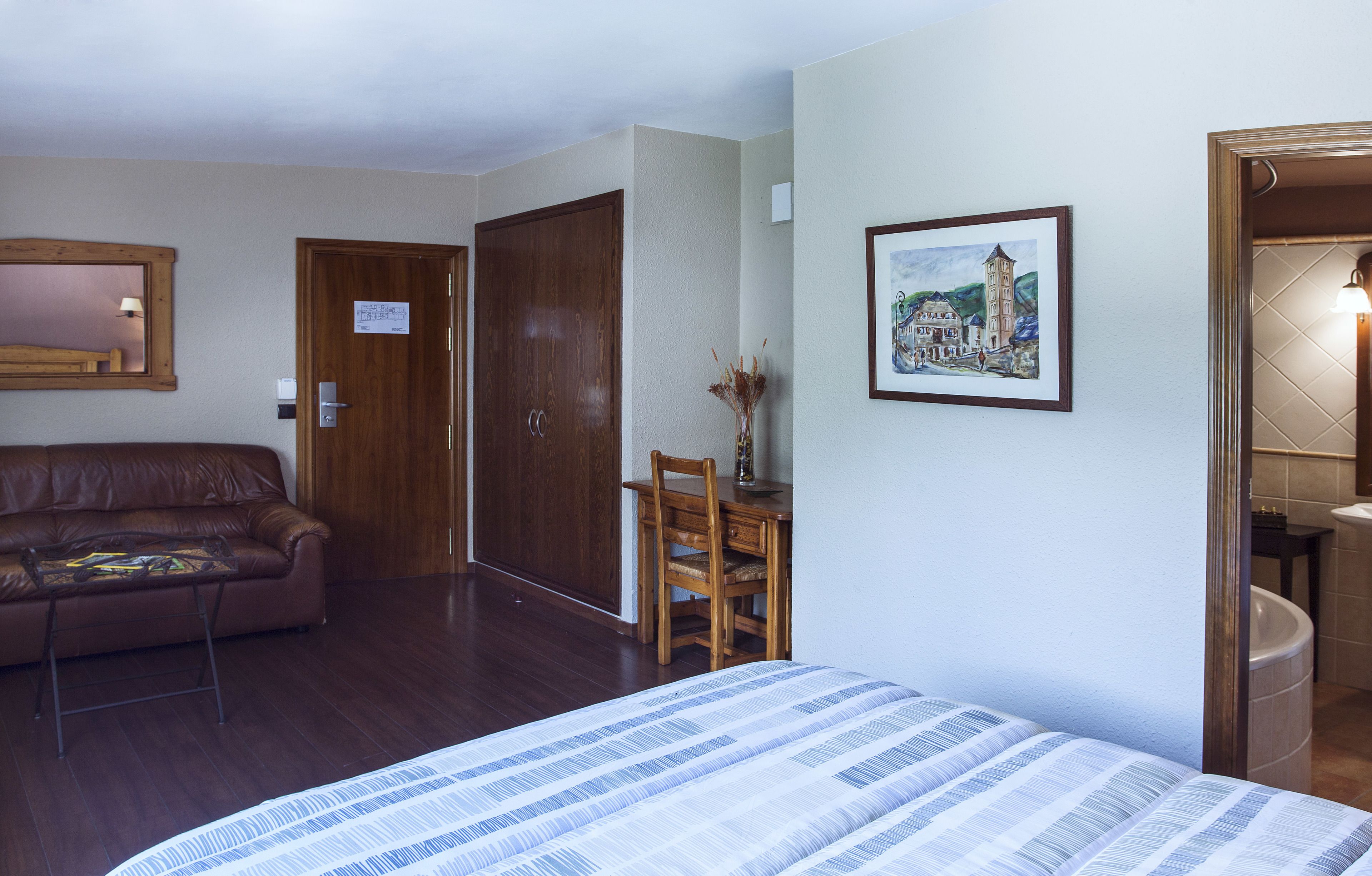 A hotel room with a bed, a wooden wardrobe, a brown leather sofa, a small table with a vase, and a framed picture on the wall.