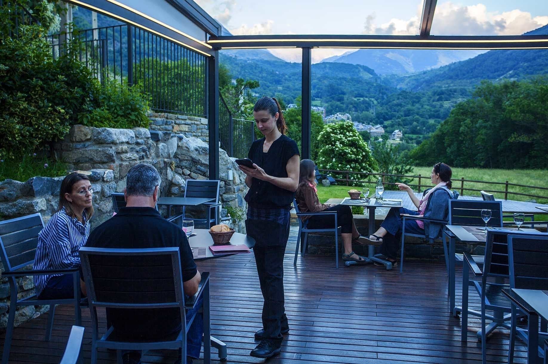 A covered outdoor seating area with stone walls, wooden flooring, and tables and chairs.