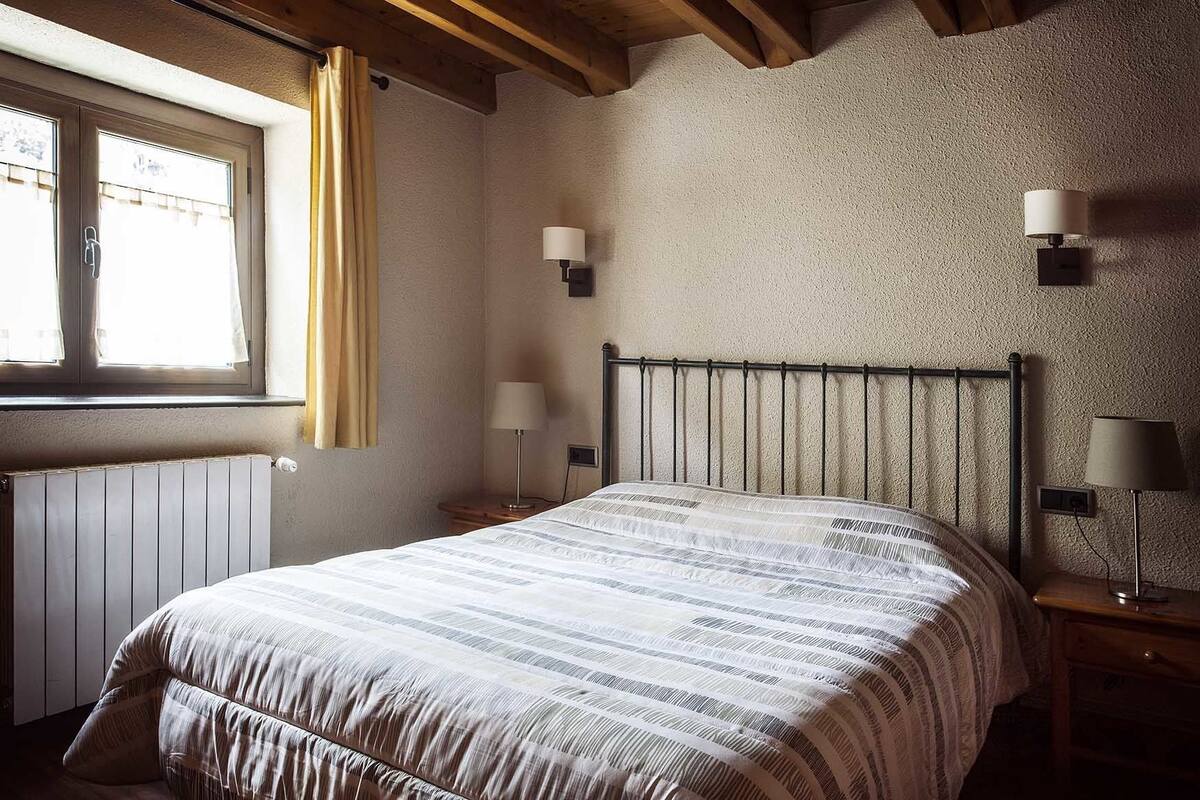 Un dormitorio con una cama, dos lámparas de noche, una ventana con cortinas y un radiador.