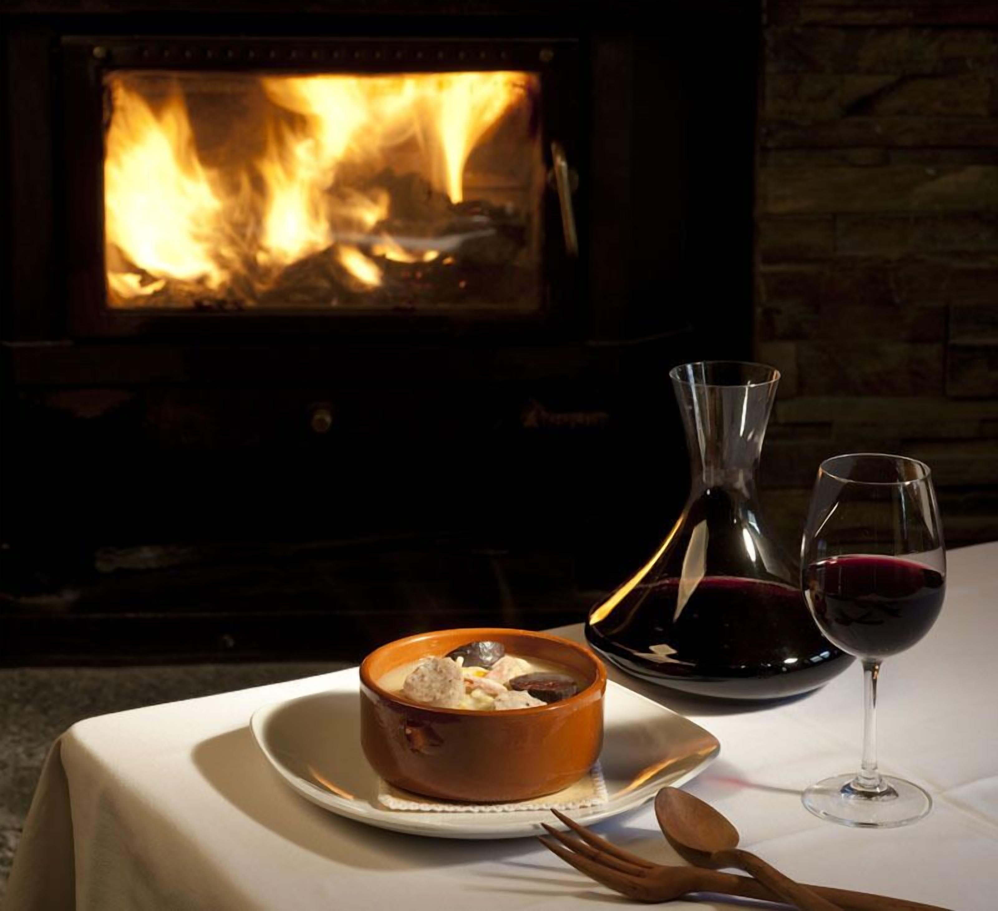 Una mesa con un bol de comida, una copa de vino y un botellero, con un hogar encendido de fondo.