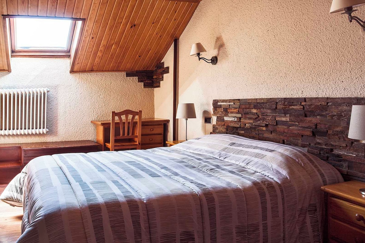 A bedroom with a sloped ceiling, a wooden headboard, a large bed with striped bedding, a wooden nightstand, a desk with a chair, a wall-mounted lamp, and a radiator.