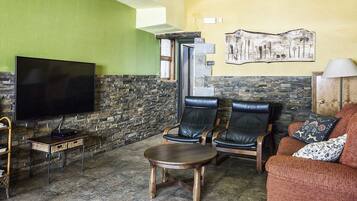 Ein Wohnzimmer mit Steinwand, einem Flachbildfernseher, zwei Ledersesseln, einem runden Holztisch und einem Sofa mit gemusterten Kissen.