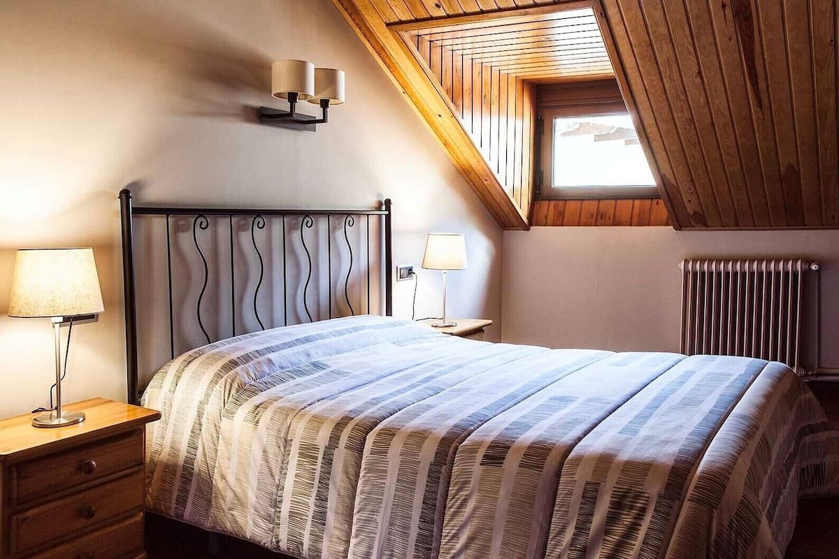 Un dormitorio con cama, lámparas de noche, cabecera de madera, mesita de noche, cajón, radiador y techo inclinado con tragaluz.