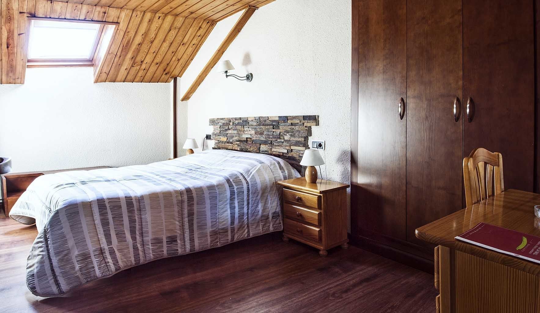 Un dormitorio con techo inclinado, cama con colcha a rayas, mesita de noche de madera con lámpara, armario de madera y una mesa de comedor con una silla.