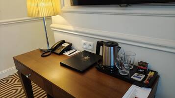 Mesa de quarto de hotel com telefone, laptop, cafeteira, copos e bandeja com petiscos.