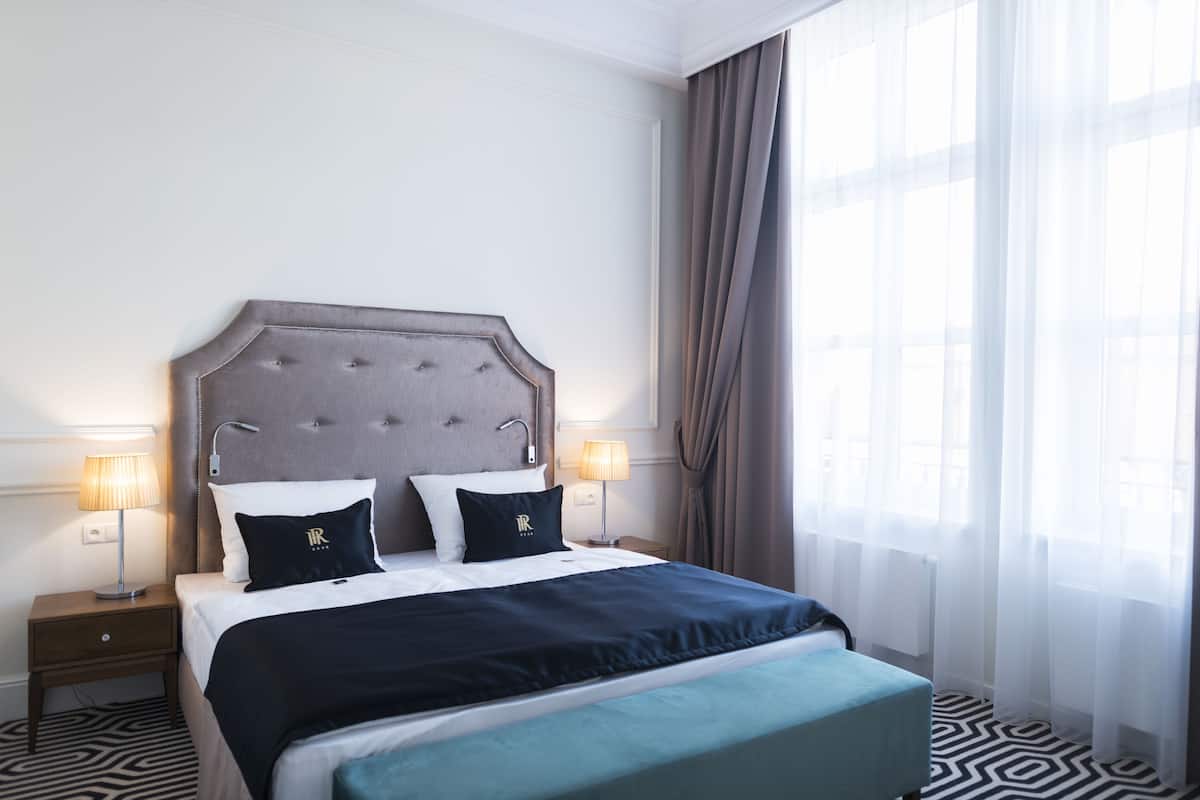 A hotel room with a large bed, two bedside tables with lamps, a headboard, and a window with curtains.