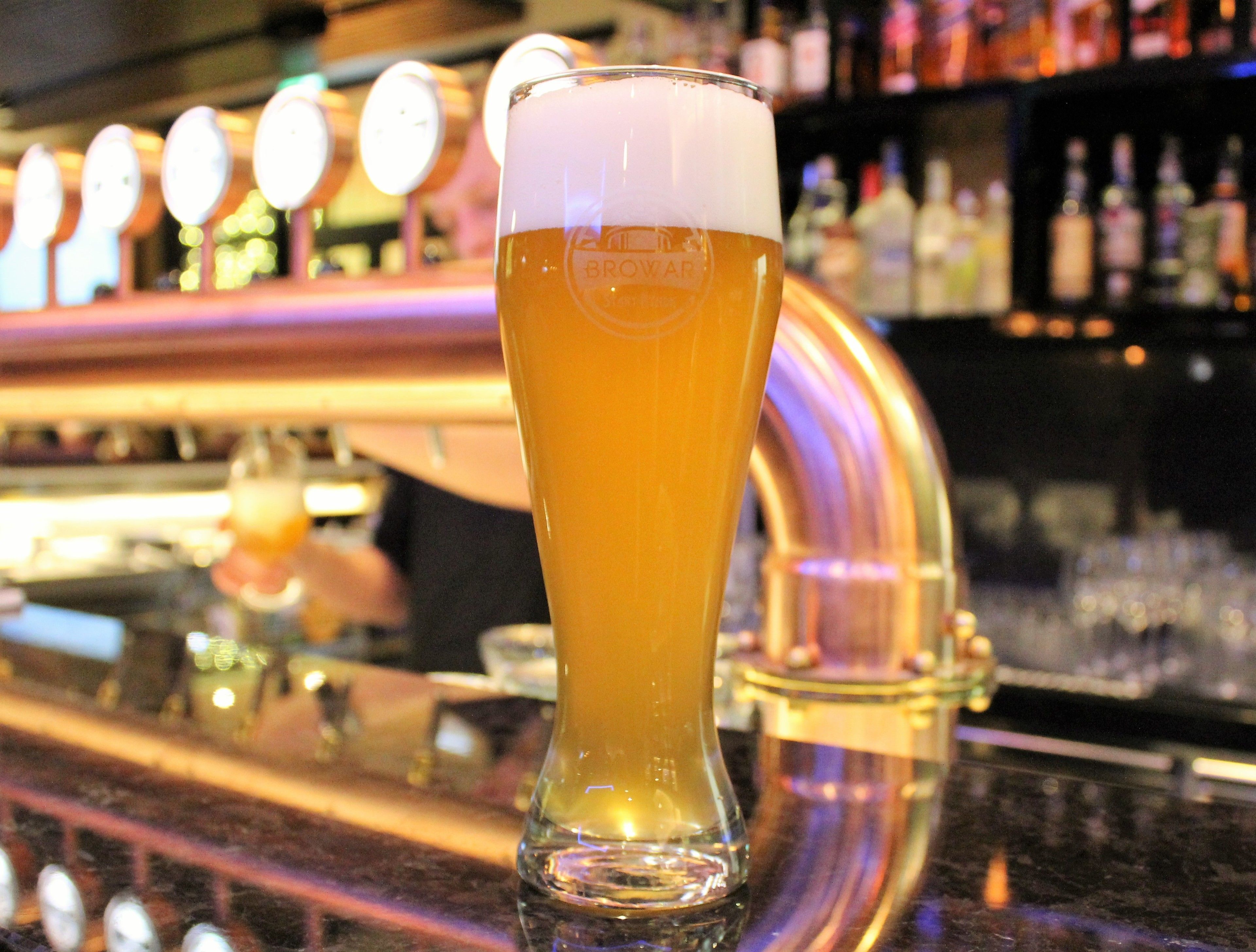 A tall glass of beer with a frothy head, placed on a bar counter with a blurred background of bar equipment and bottles.