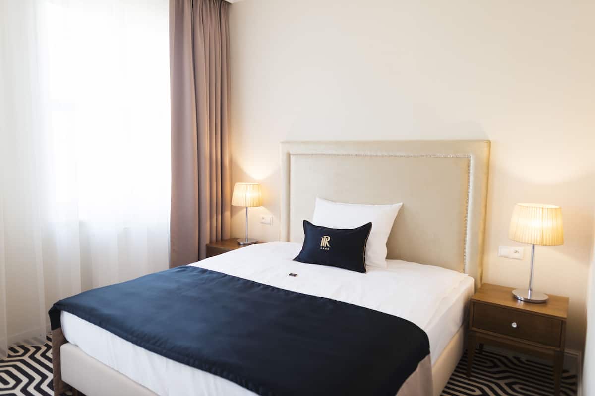 A hotel room with a large bed, two bedside tables with lamps, a headboard, and a window with curtains.