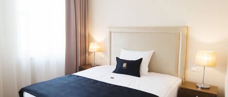 A hotel room with a large bed, two bedside tables with lamps, a headboard, and a window with curtains.