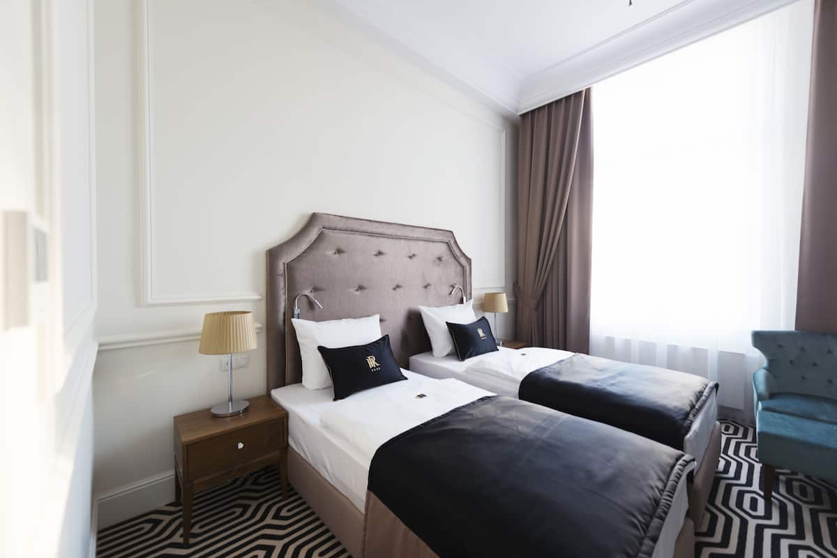 A hotel room with two beds, a nightstand with a lamp, a chair, and a window with curtains.
