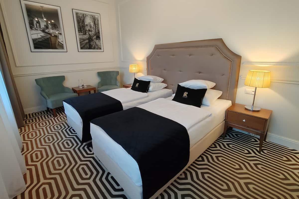 A hotel room with two beds, a patterned floor, a green armchair, and two framed pictures on the wall.