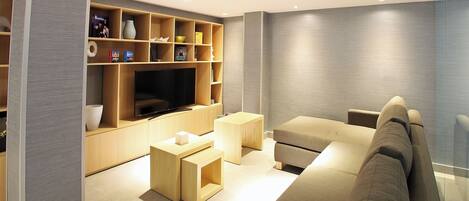 Een moderne woonkamer met een grote flatscreen televisie, houten wandplanken, een beige bank en een glazen scheidingswand.