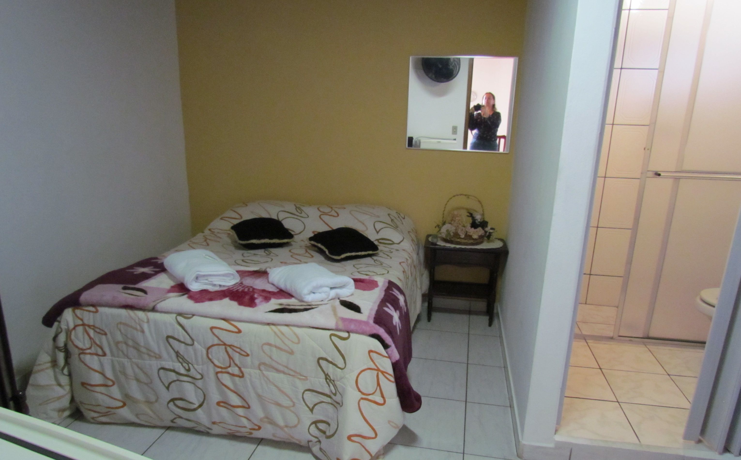 A small, neatly made bed with a floral bedspread and two pillows, a bedside table with a lamp and a decorative item, and a mirror on the wall.