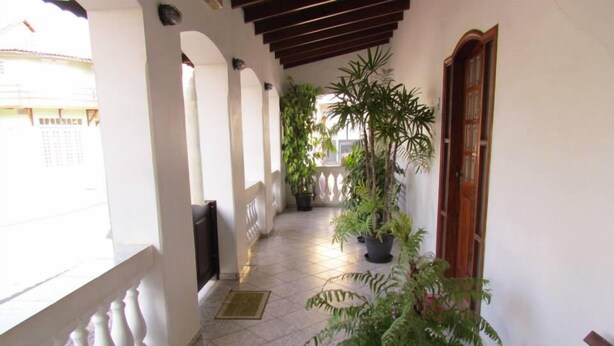 A covered corridor with tiled flooring, white walls, and wooden beams. There are potted plants along the corridor, and a door with a wooden frame on the right. - Hotel Pousada Alvorada (Brotas)