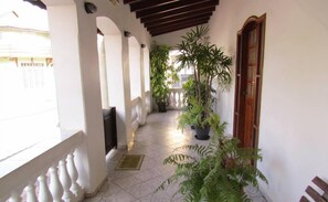 A covered corridor with tiled flooring, white walls, and wooden beams. There are potted plants along the corridor, and a door with a wooden frame on the right. - Hotel Pousada Alvorada (Brotas)